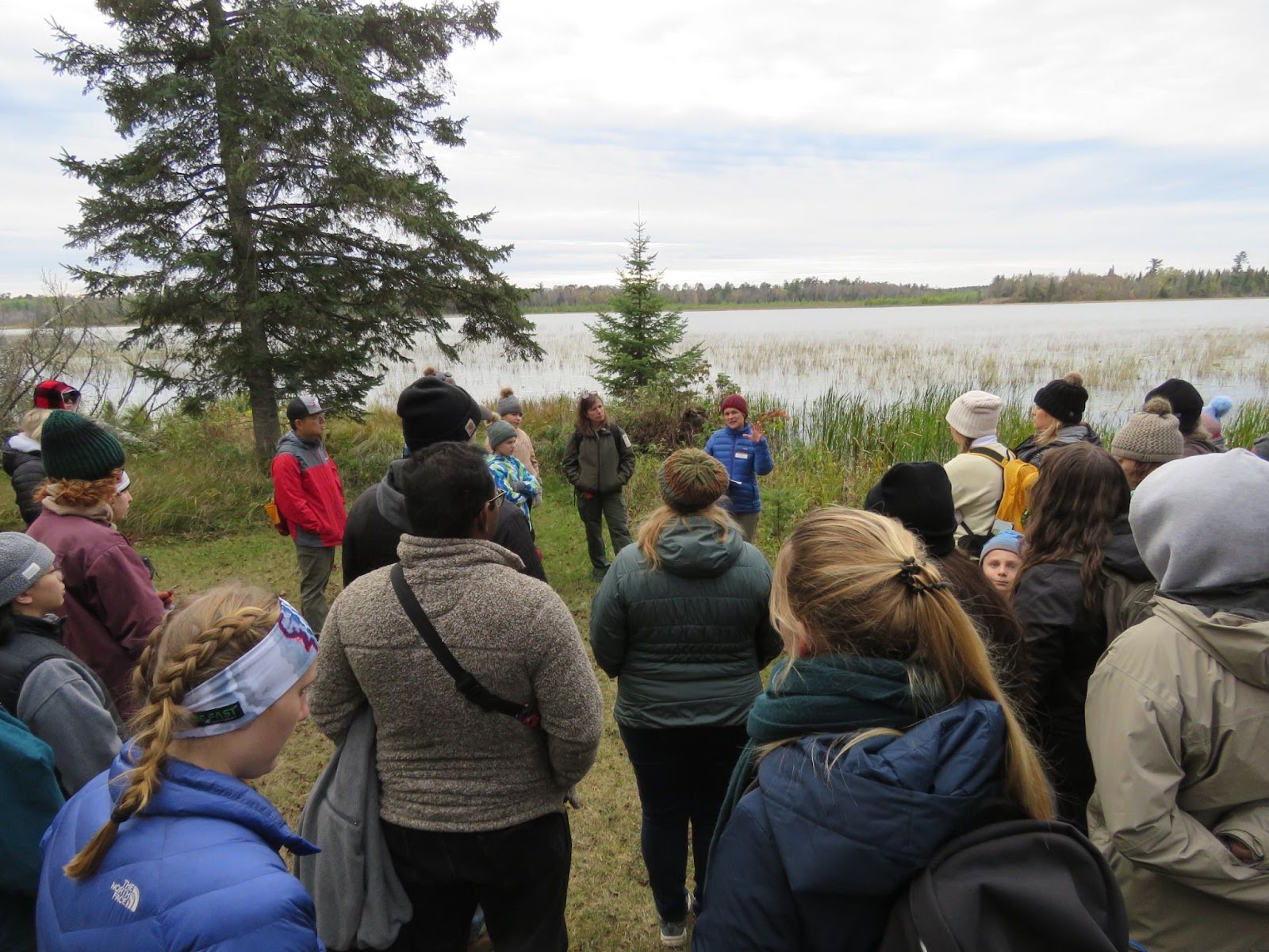 A group of people listening to Emily Schilling and and an Itasca State Park naturalist at Bear Paw Point at the edge of Lake Itasca