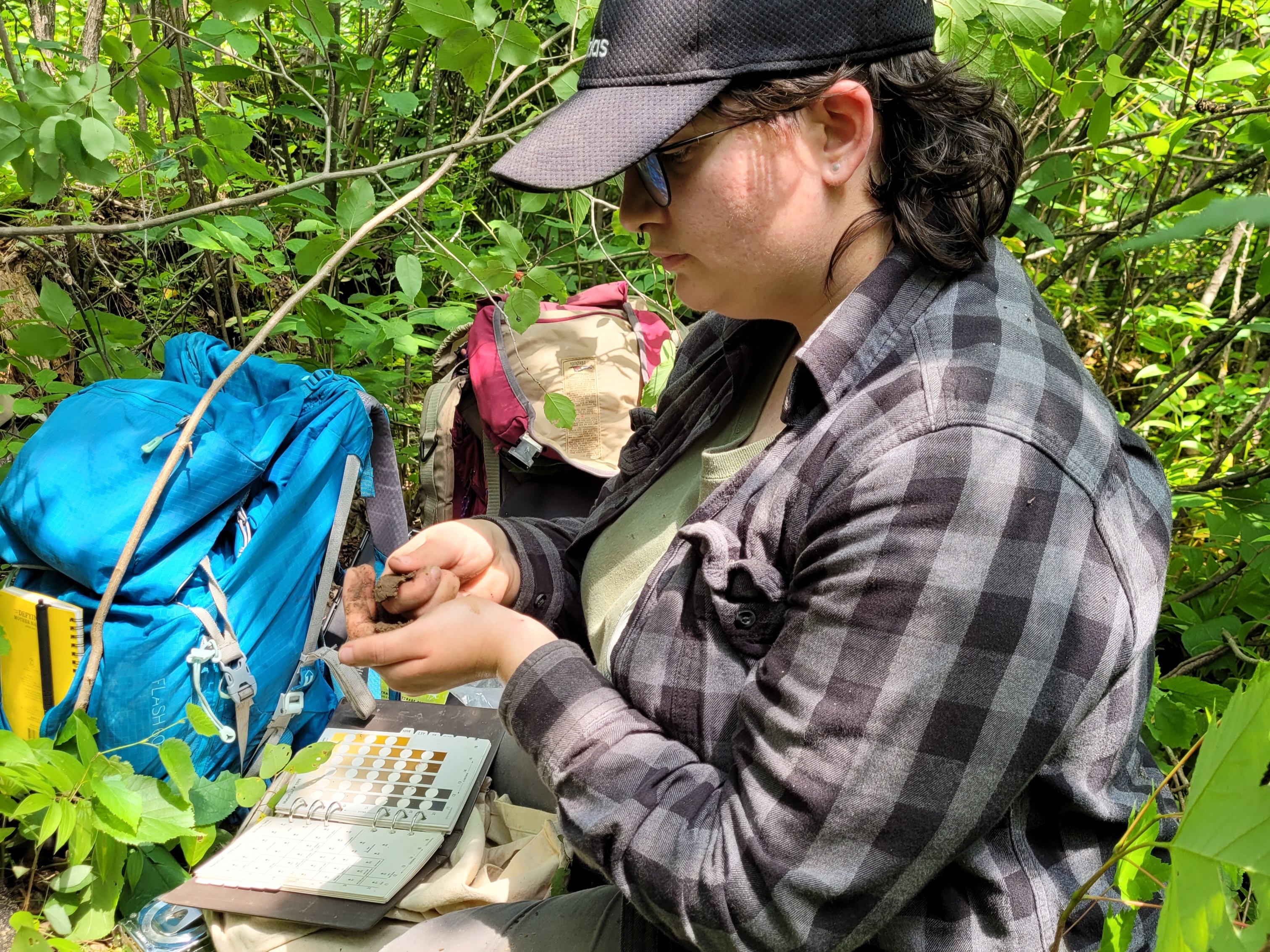 A researcher sitting on the ground in the forest, looking at soil in her hand