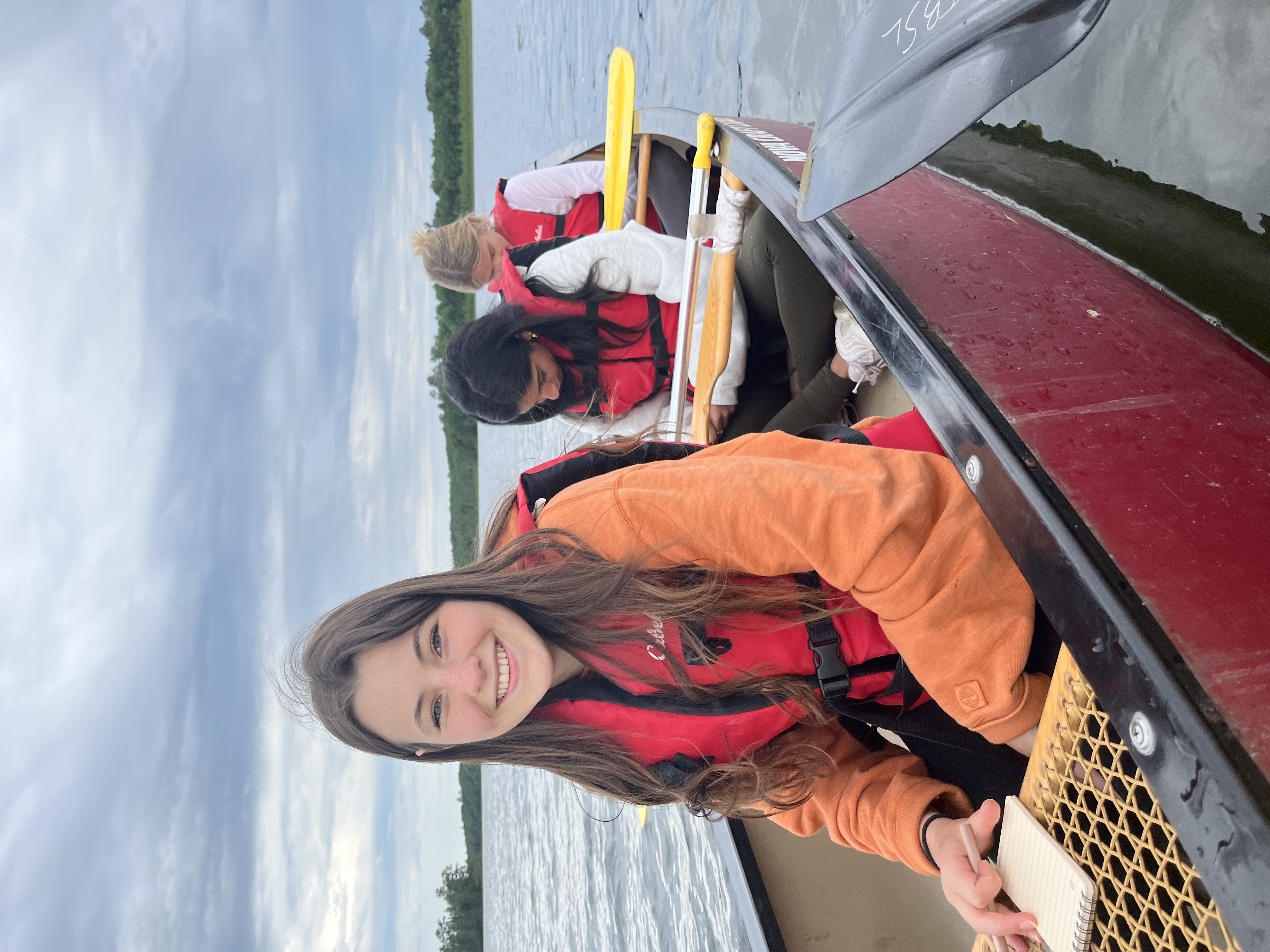 Three NOL students in a canoe on Lake Itasca. The lead student is smiling at the camera, the other two students are writing in notebooks.