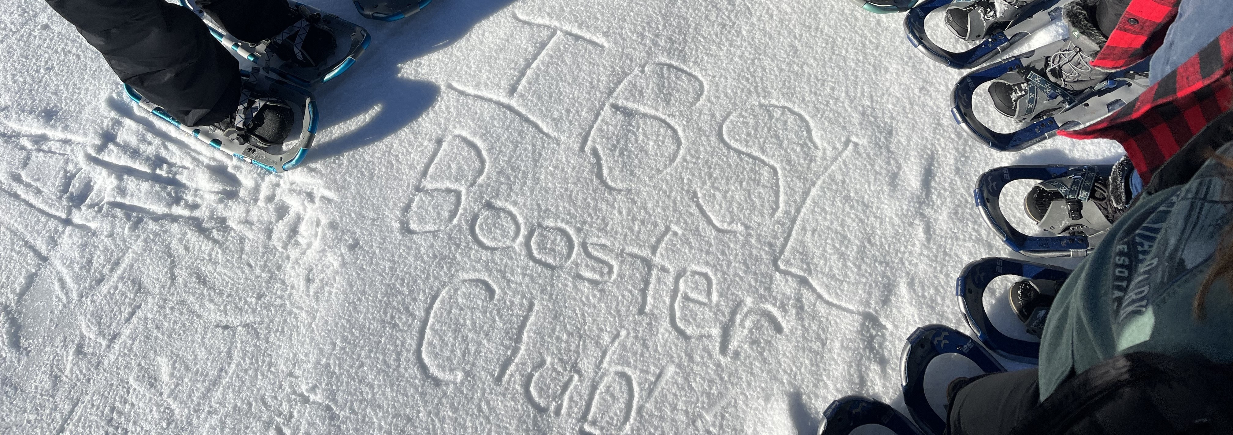 Only snowshoes visible from a group of people standing around the words "IBSL Booster Club" written in the snow.