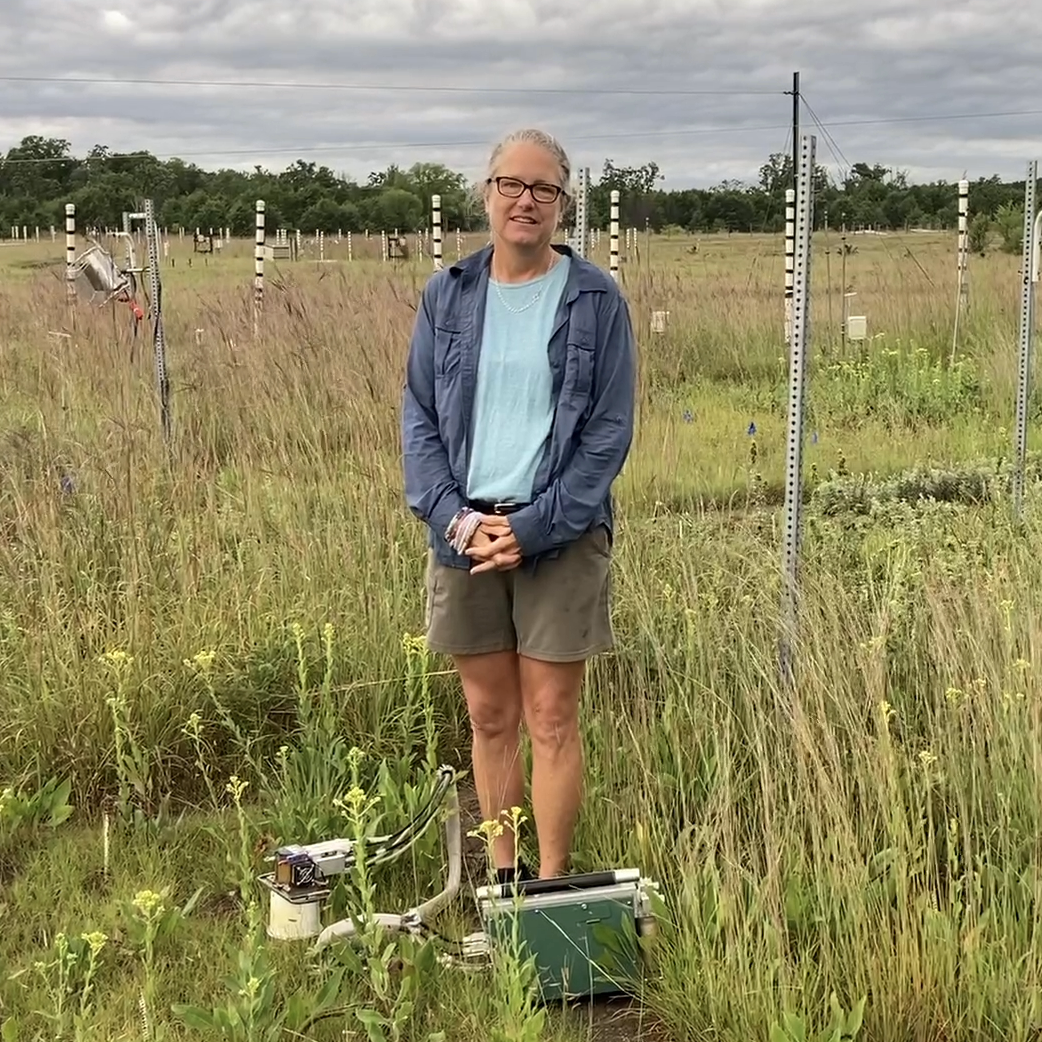 a white woman with glasses standing in a field with scientific equipment around her