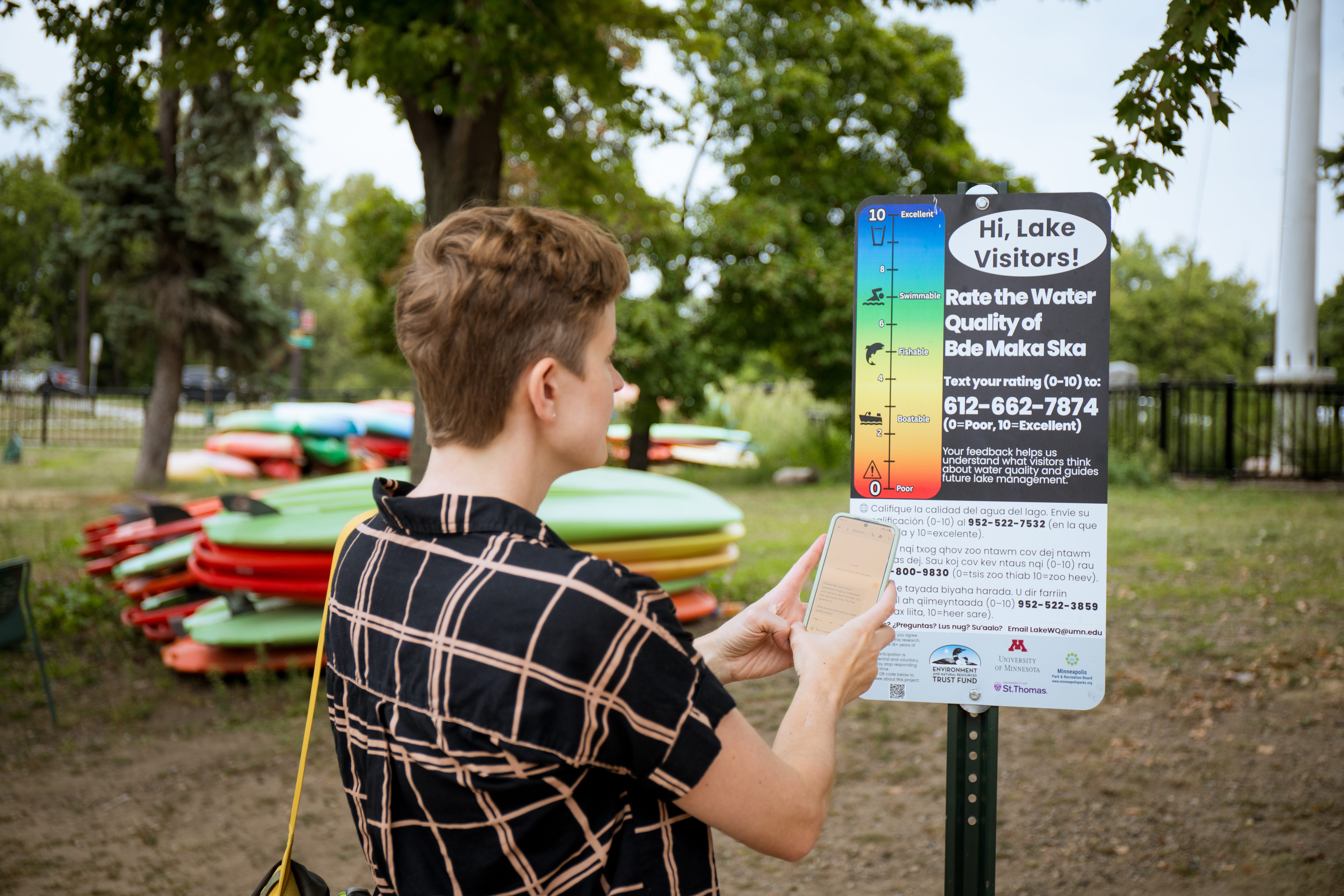 A person types on their phone in front of a sign that reads "Hi Lake Visitors! Rate the Water Quality of Bde Maka Ska."