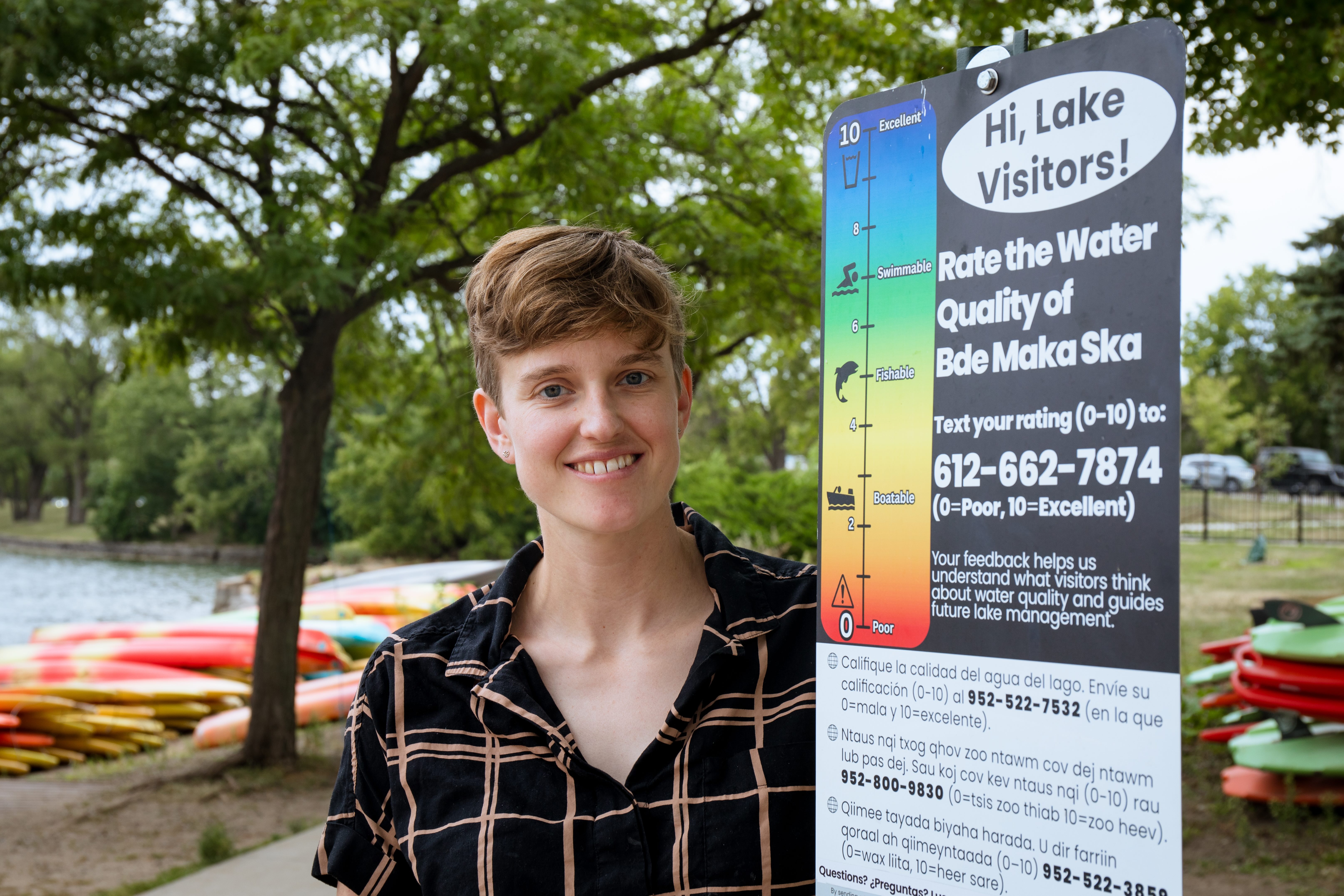 A person with short hair, wearing a black and brown striped shirt, stands next to a sign that reads "Hi Lake Visitors! Rate the Water Quality of Bde Maka Ska"