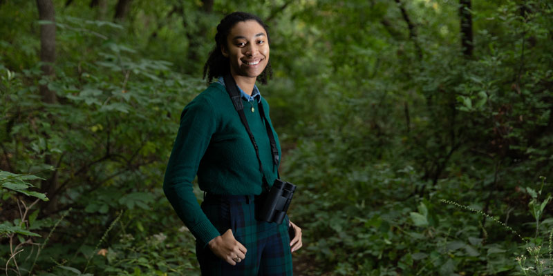 Simone Maddox stands in the woods with binoculars and looks for birds 