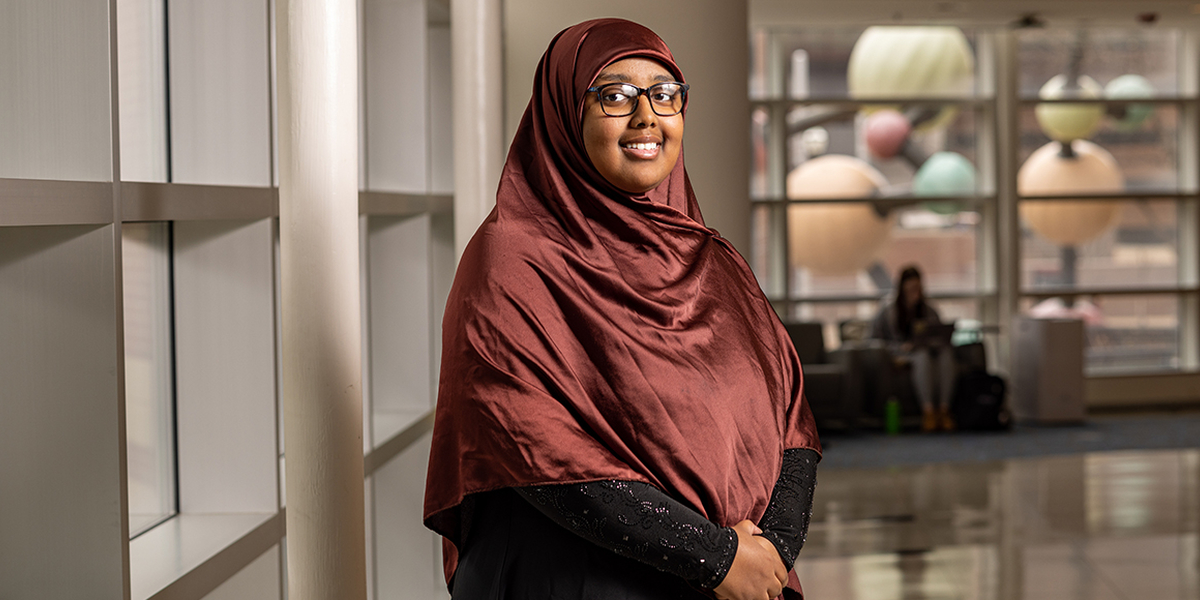 Idil Abdi wears a maroon headscarf and smiles in the hallway with windows and the MCB molecule structure in background.