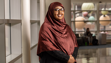 Idil Abdi wears a maroon headscarf and smiles in the hallway with windows and the MCB molecule structure in background.