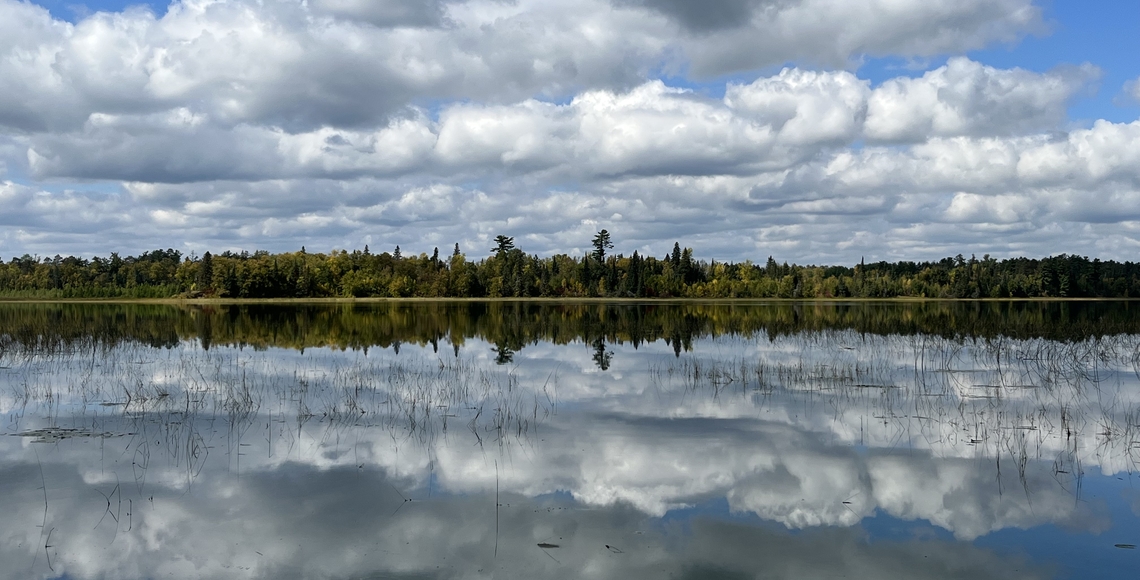 Cloudy sky reflected on the lake and treeline of the western shore of Lake Itasca, as seen from the Itasca Biological Station
